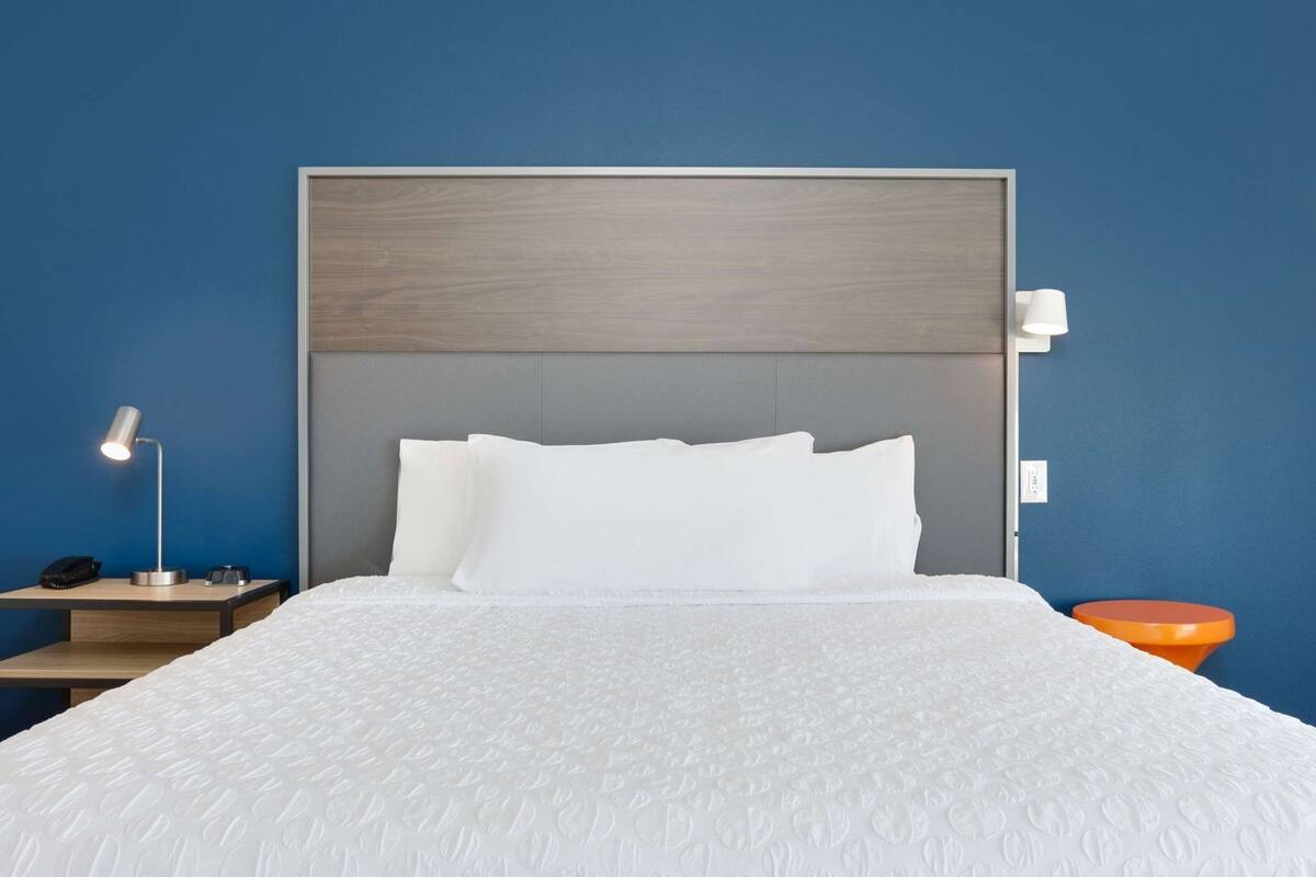 A hotel room with a large bed, a bedside lamp, a nightstand, and a small orange stool.