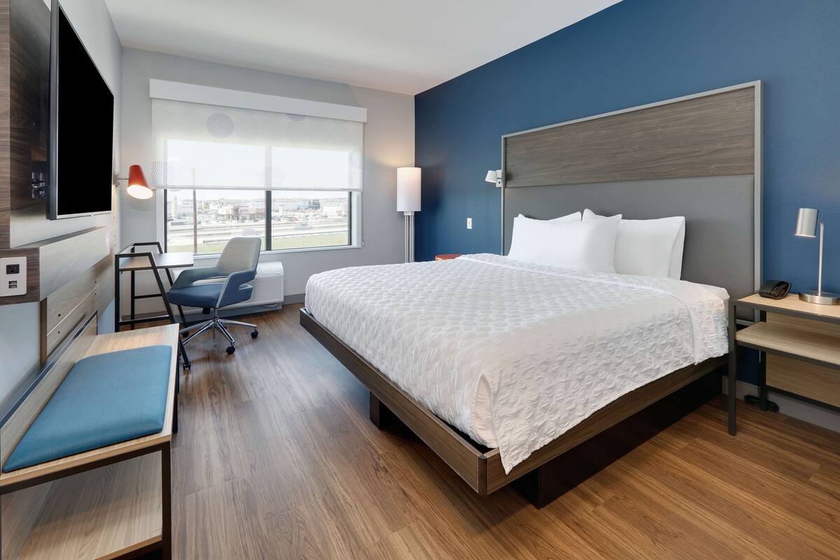Habitación de hotel moderna con una cama grande, un escritorio, una silla, un televisor montado en la pared y una ventana con vistas a la ciudad.