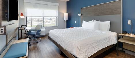 A modern hotel room with a large bed, a desk, a chair, a TV mounted on the wall, and a window with a city view.