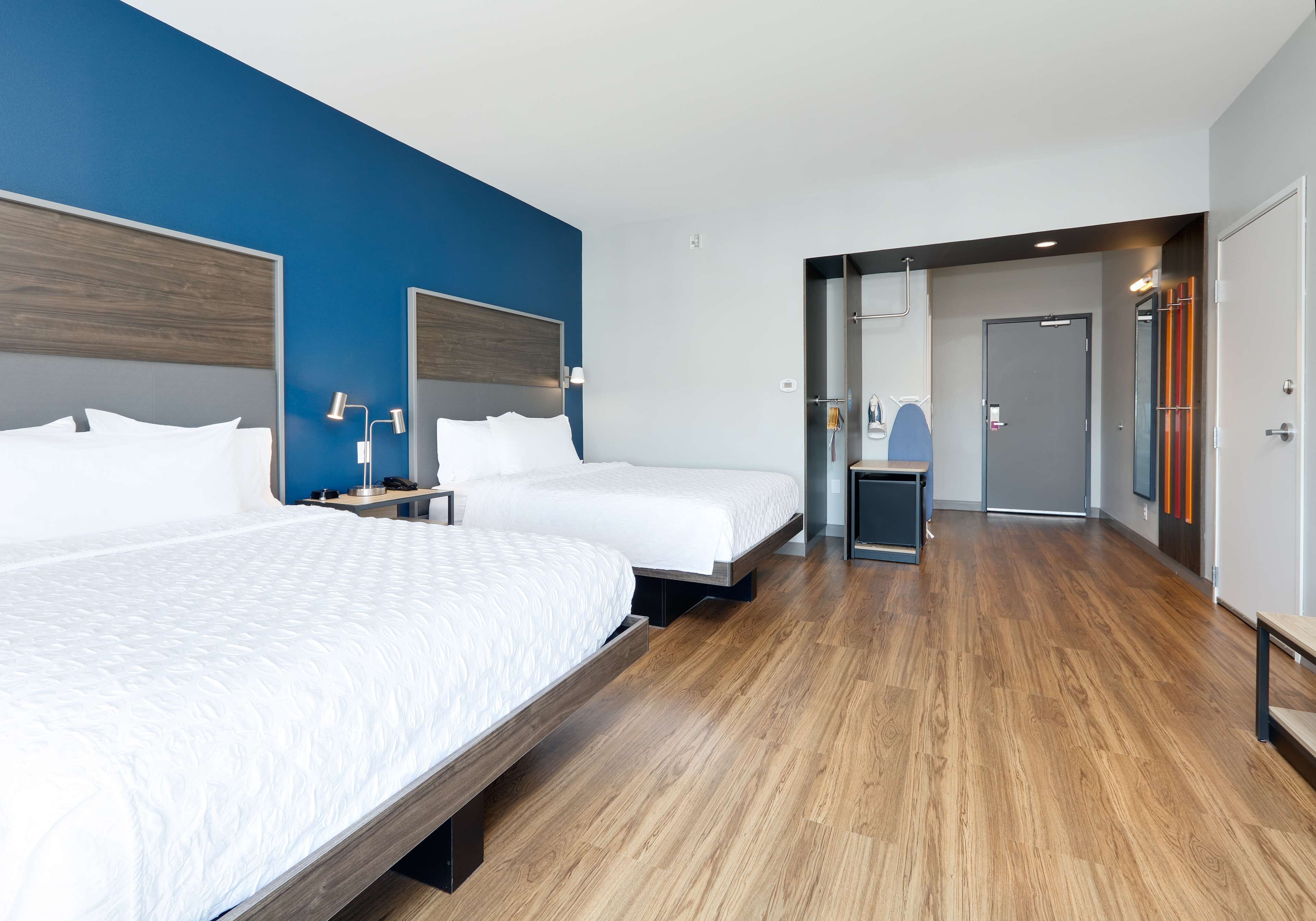 A modern hotel room with two beds, a wooden floor, and a bathroom visible through an open door.