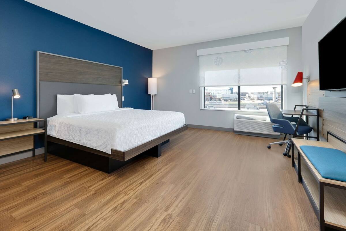 A modern hotel room with a bed, desk, chair, and large window with city view.
