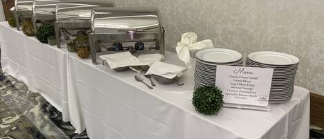 A buffet setup with various food items and utensils, a menu sign, and a decorative plant.