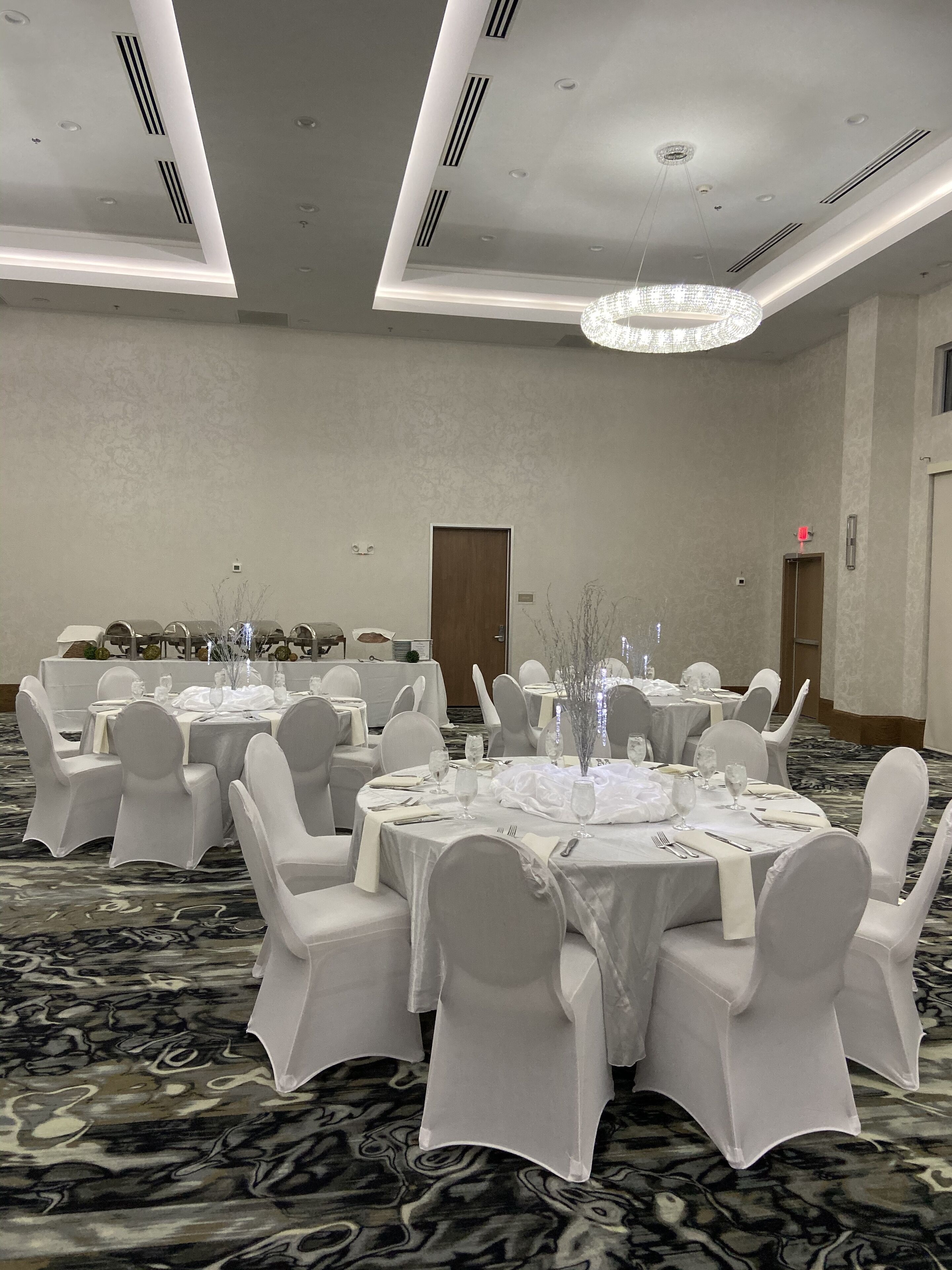 Un salón de banquetes con mesas redondas dispuestas para comer, sillas blancas y una araña de cristal.