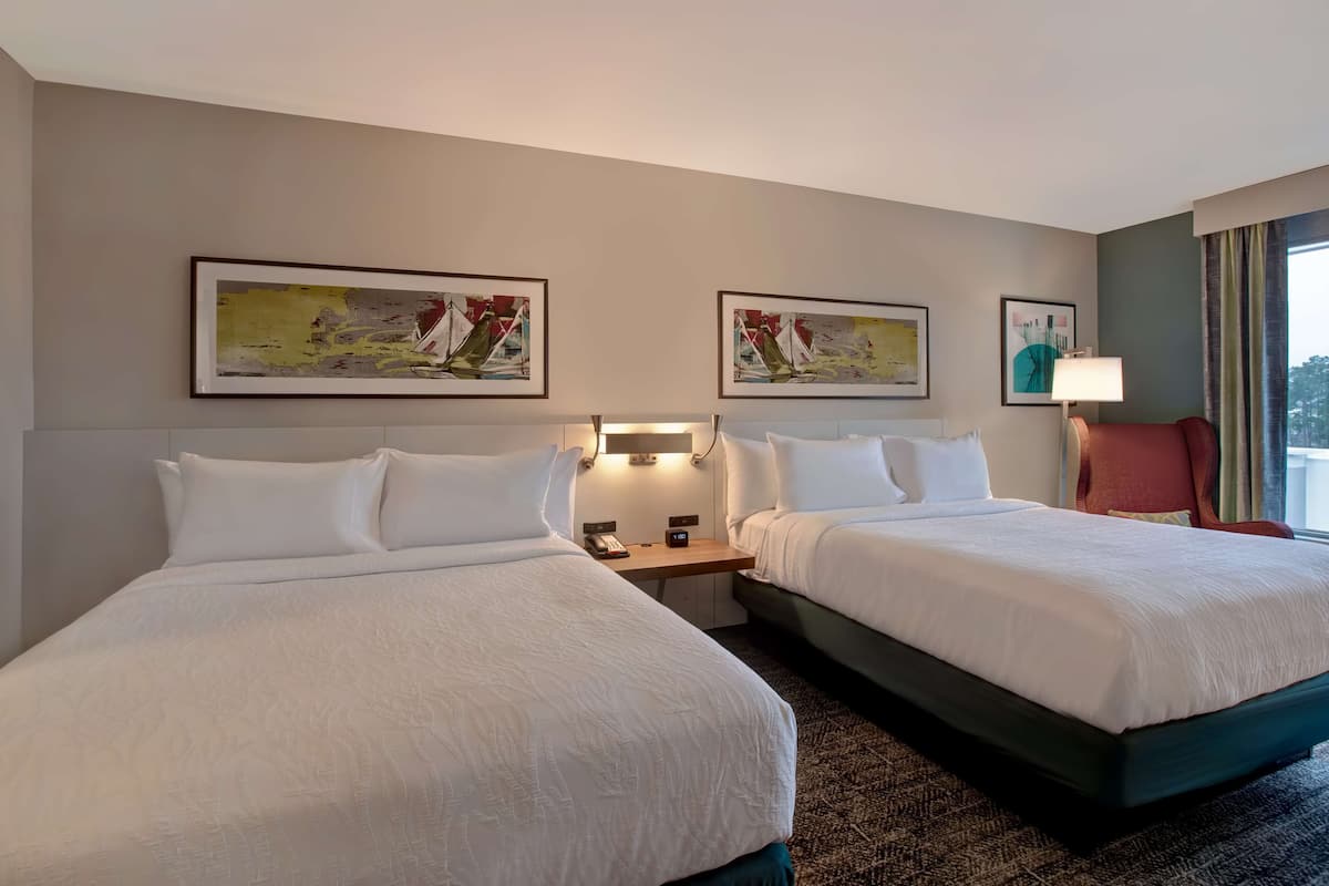 A hotel room with two beds, a nightstand with a lamp, and artwork on the walls.