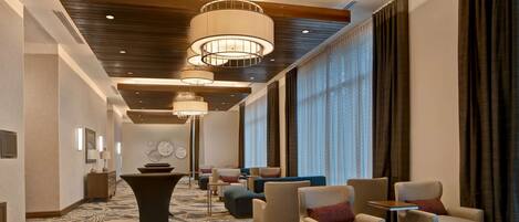Um saguão de hotel com carpete estampado, área de estar com poltronas e sofás, uma mesa de centro e iluminação pendente.