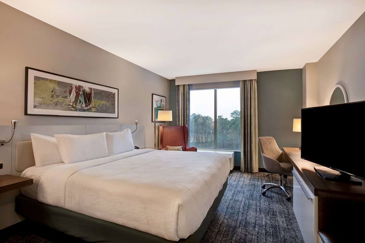 A hotel room with a large bed, a desk with a flat-screen TV, a chair, a lamp, and a window with a view of trees.