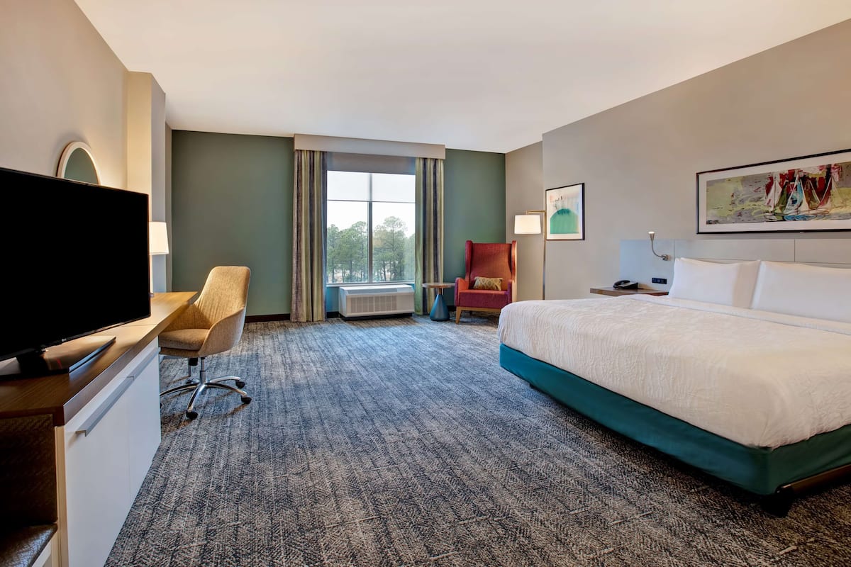 A hotel room with a bed, a desk with a TV, a chair, a window with curtains, and a painting on the wall.