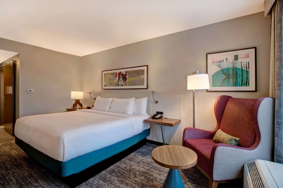 A hotel room with a large bed, bedside tables, a chair, a round table, and artwork on the walls.