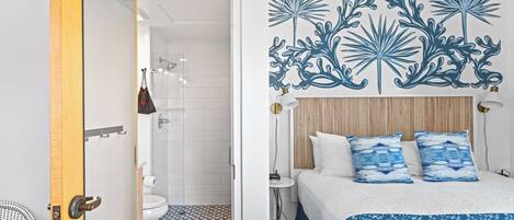 Habitación de hotel con cama, mesita de noche, una obra de arte en la pared y baño con ducha e inodoro.