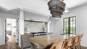 Ett kök med en stor köksö, flätade stolar, en stänkskydd av sten och en hängande lampa.