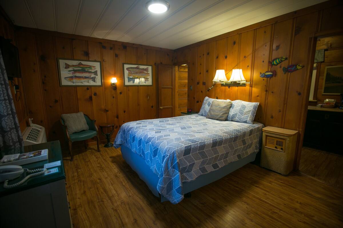 A wooden-paneled room with a bed, a chair, a desk with a phone, and wall-mounted lamps.