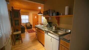 A compact kitchen with wooden cabinets, a built-in dishwasher, and a microwave. There is a dining area with a chair and a table, and a wooden door leading to another room.
