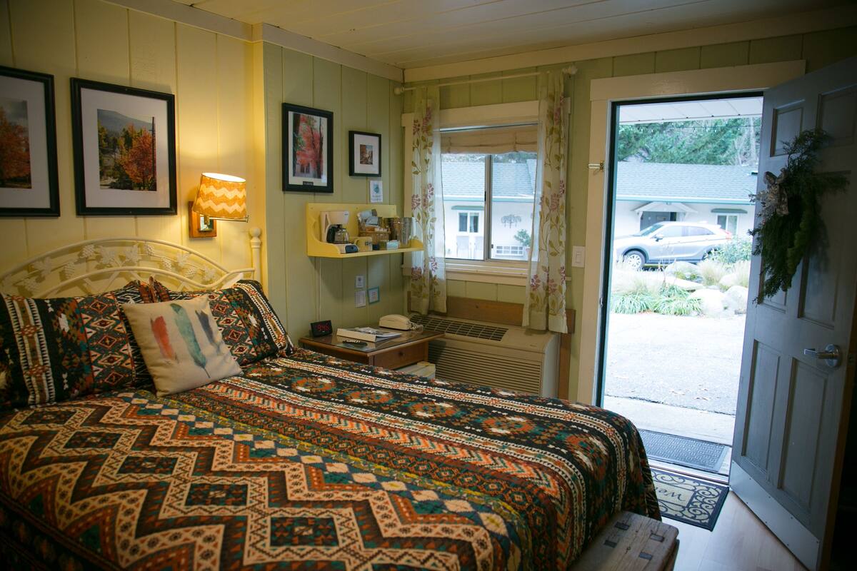 A bedroom with a large bed, a bedside table with a lamp, a window with curtains, and a door with a wreath.