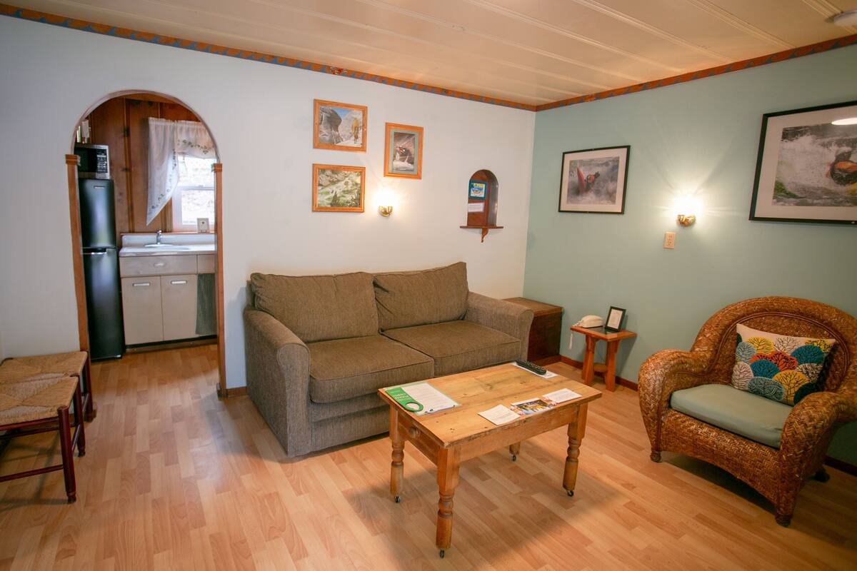 A living room with a sofa, armchair, wooden coffee table, and a small side table. There is a kitchen area with a refrigerator and microwave visible through an archway.