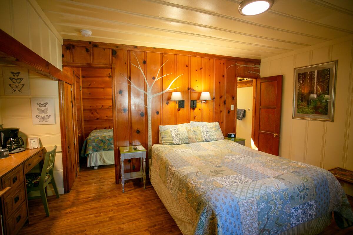 A cozy bedroom with a wooden floor, a bed with a floral quilt, a desk with a chair, and a nightstand with lamps.