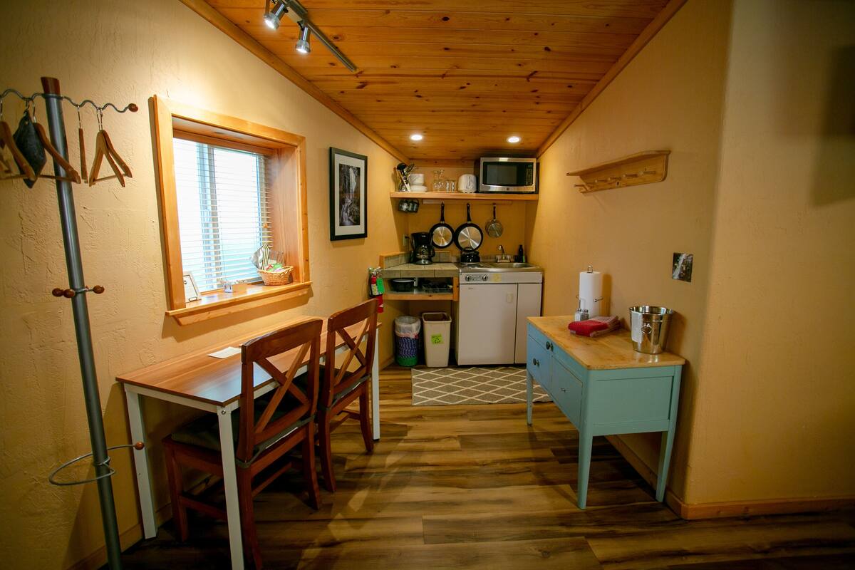 A compact kitchen with wooden flooring, a dining table with chairs, a microwave, and a small refrigerator.