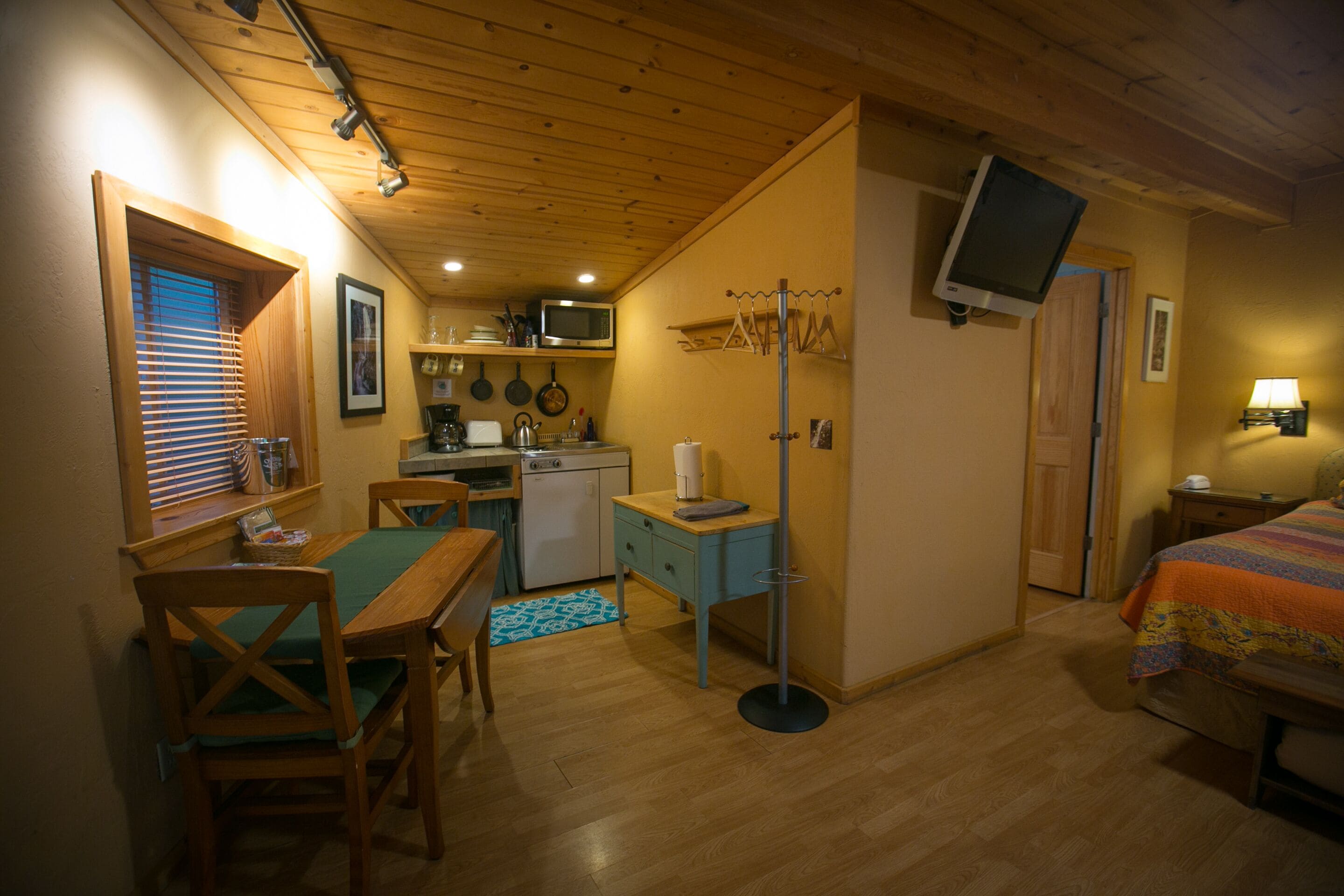 A compact kitchen with wooden cabinets, a microwave, and a sink. There is a dining table with chairs and a blue rug. A bed with an orange blanket is visible in the corner.