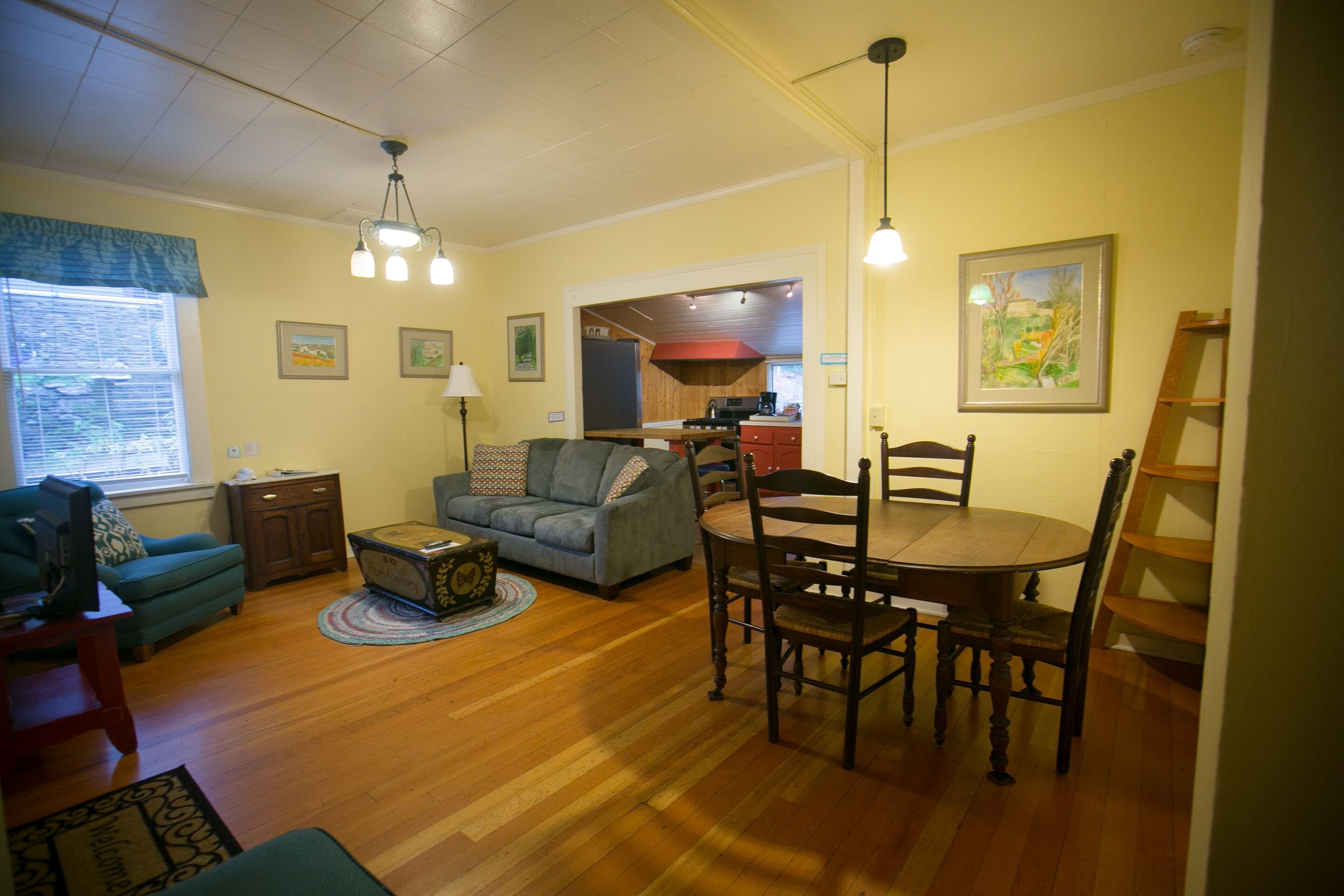 A living room with a sofa, a coffee table, and a dining table with chairs. There is a window with blinds, a lamp, and framed pictures on the wall.