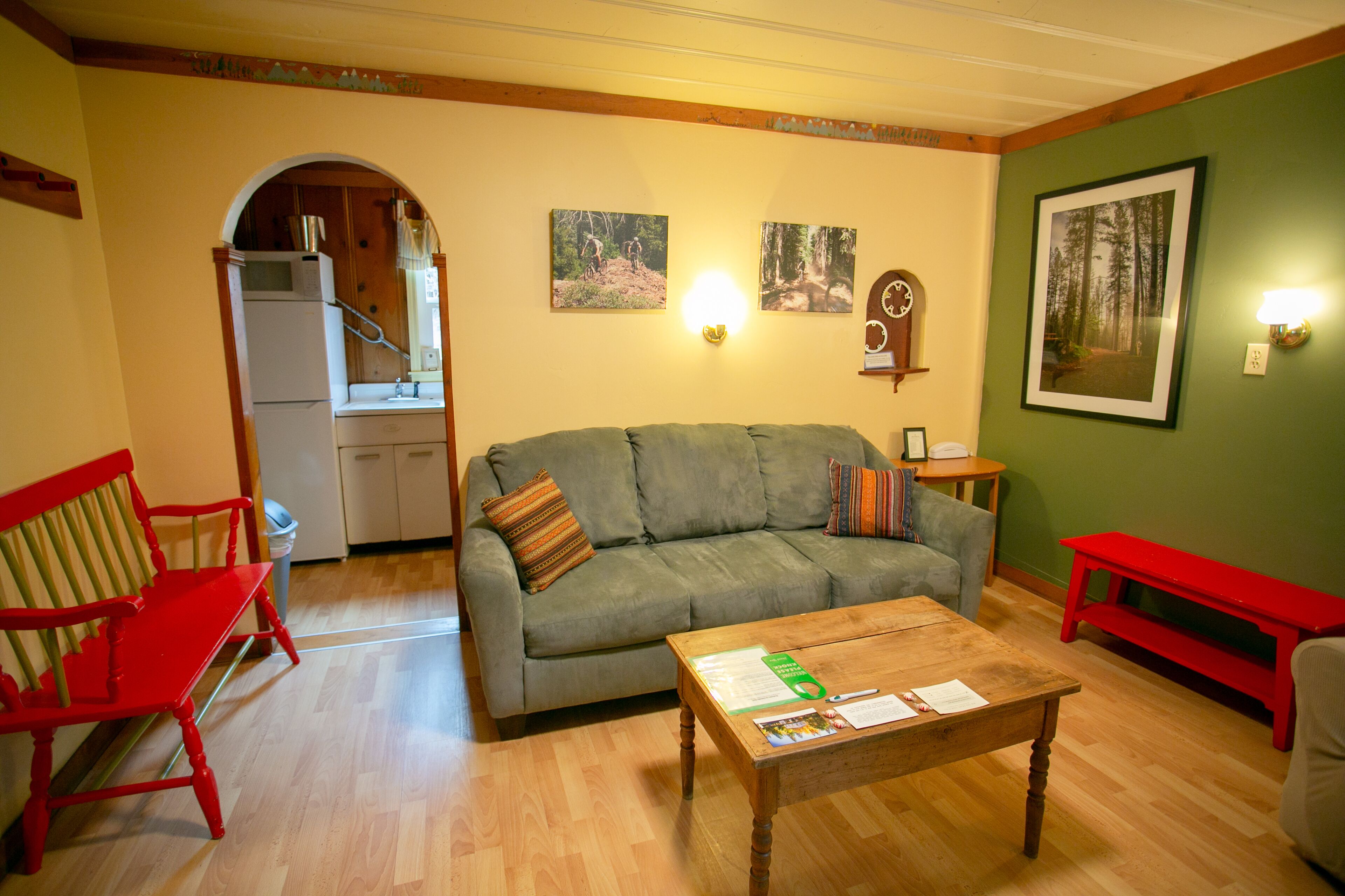Una sala de estar con un sofá, una mesa de centro, un banco rojo, una silla roja, una zona de cocina con un refrigerador y una escalera.