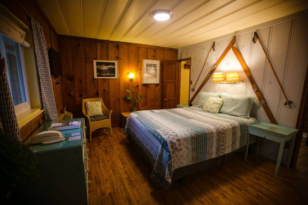 A cozy bedroom with a wooden floor, a bed with a patterned blanket, a nightstand, a chair, and a desk with a telephone.
