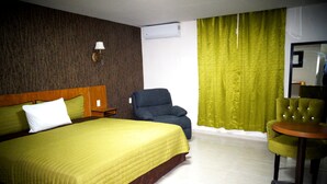 A hotel room with a large bed, two chairs, a small table, a nightstand, a wall-mounted lamp, and a window with curtains.