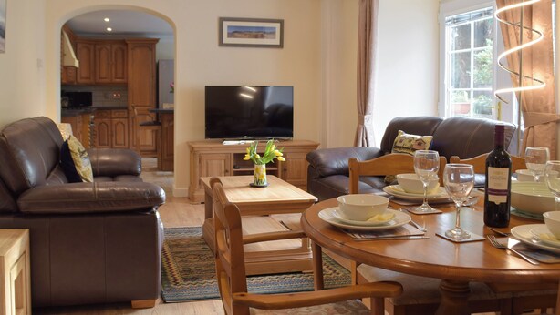 A dining area with a wooden table set for four, a television, and a kitchen in the background. - Victoria House - UK13380 (Llansteffan, near Carmarthen)