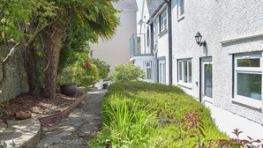 A narrow pathway flanked by lush greenery and a white building with multiple windows and a satellite dish. - Victoria House - UK13380 (Llansteffan, near Carmarthen)
