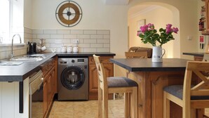 A modern kitchen with a built-in washer and dryer, wooden bar stools, a clock on the wall, and a vase with flowers on the counter. - Victoria House - UK13380 (Llansteffan, near Carmarthen)