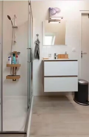 Une salle de bain avec une douche à parois de verre, un meuble-lavabo blanc avec un miroir et une petite étagère en bois.