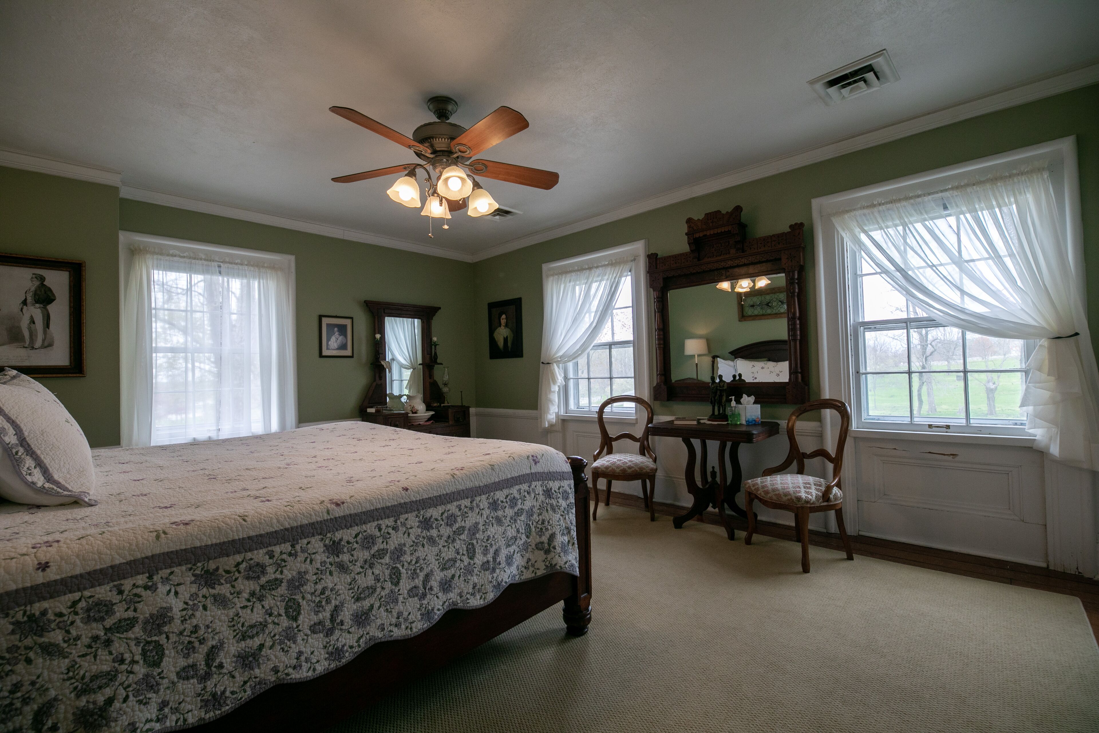 A bedroom with a bed, a desk, two chairs, a ceiling fan, and large windows with curtains.