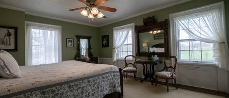 A bedroom with a bed, a desk, two chairs, a ceiling fan, and large windows with curtains.