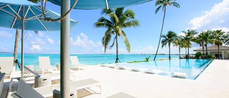 A poolside area with lounge chairs and umbrellas, palm trees, and a clear view of the ocean.