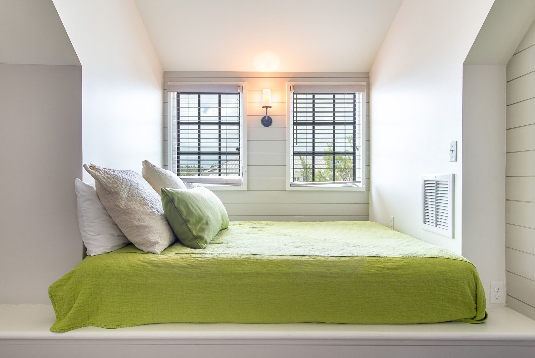 A bedroom with a sloped ceiling, two windows with shutters, a bed with a green bedspread, and a wall-mounted light fixture.