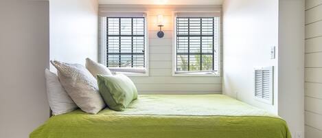 A bedroom with a sloped ceiling, two windows with shutters, a bed with a green bedspread, and a wall-mounted light fixture.