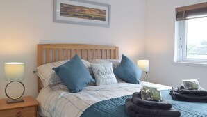 A neatly made bed with blue and white bedding, two bedside lamps, a framed picture on the wall, and a window with blinds. - 2 bedroom accommodation in Llansteffan, near Carmarthen (Llansteffan, near Carmarthen)