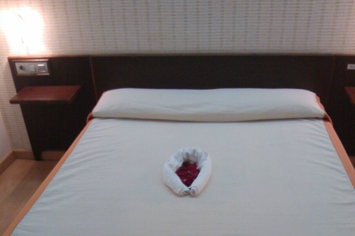 A neatly made bed with a folded towel in the center, a wooden nightstand to the left, and a headboard against a patterned wall.