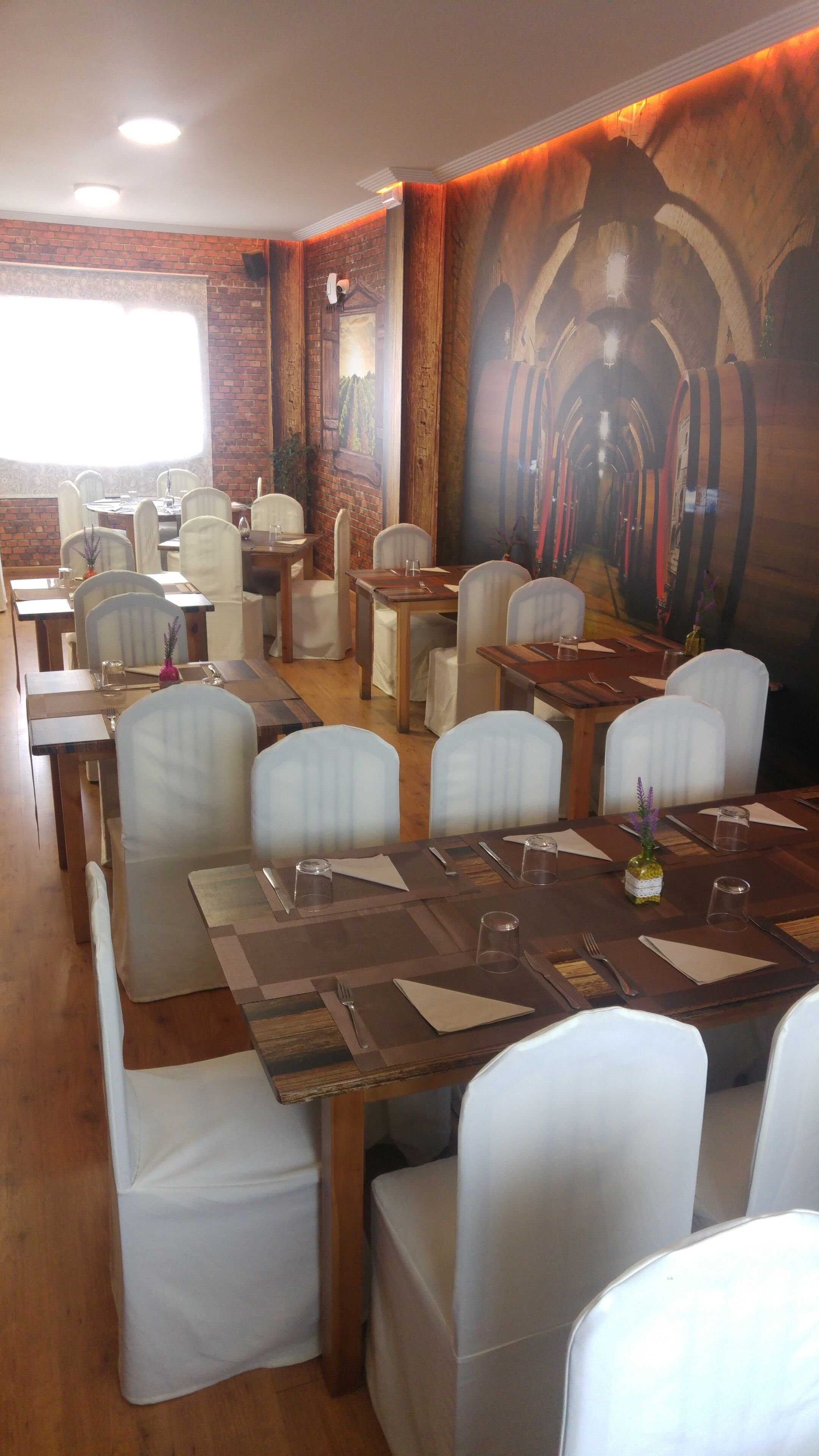 A restaurant with wooden tables and white chairs, brick walls, and a mural of barrels.
