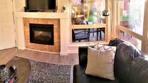 Una sala de estar con chimenea, un televisor montado encima, un sofá con cojines y vistas a un patio exterior a través de una ventana.