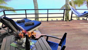 Une terrasse en bois avec un fauteuil de relaxation et un hamac, donnant sur une plage bordée de palmiers et sur l’océan.