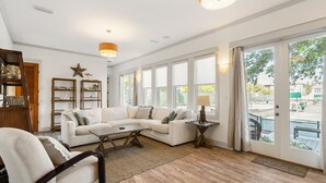 Un salon spacieux avec un grand canapé d’angle blanc, une table basse et un plancher en bois. De grandes fenêtres sont agrémentées de rideaux, et on y trouve un meuble en bois ainsi qu’une décoration en forme d’étoile sur le mur.