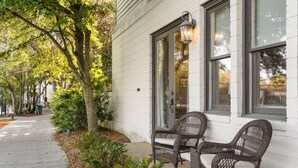 Une terrasse en bois, avec des chaises en osier et une lanterne, entourée d’arbres et d’un bâtiment blanc.