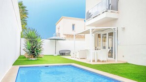 Une piscine avec un bord carrelé, un petit espace gazonné, des meubles d’extérieur blancs et un bâtiment doté d’un balcon.