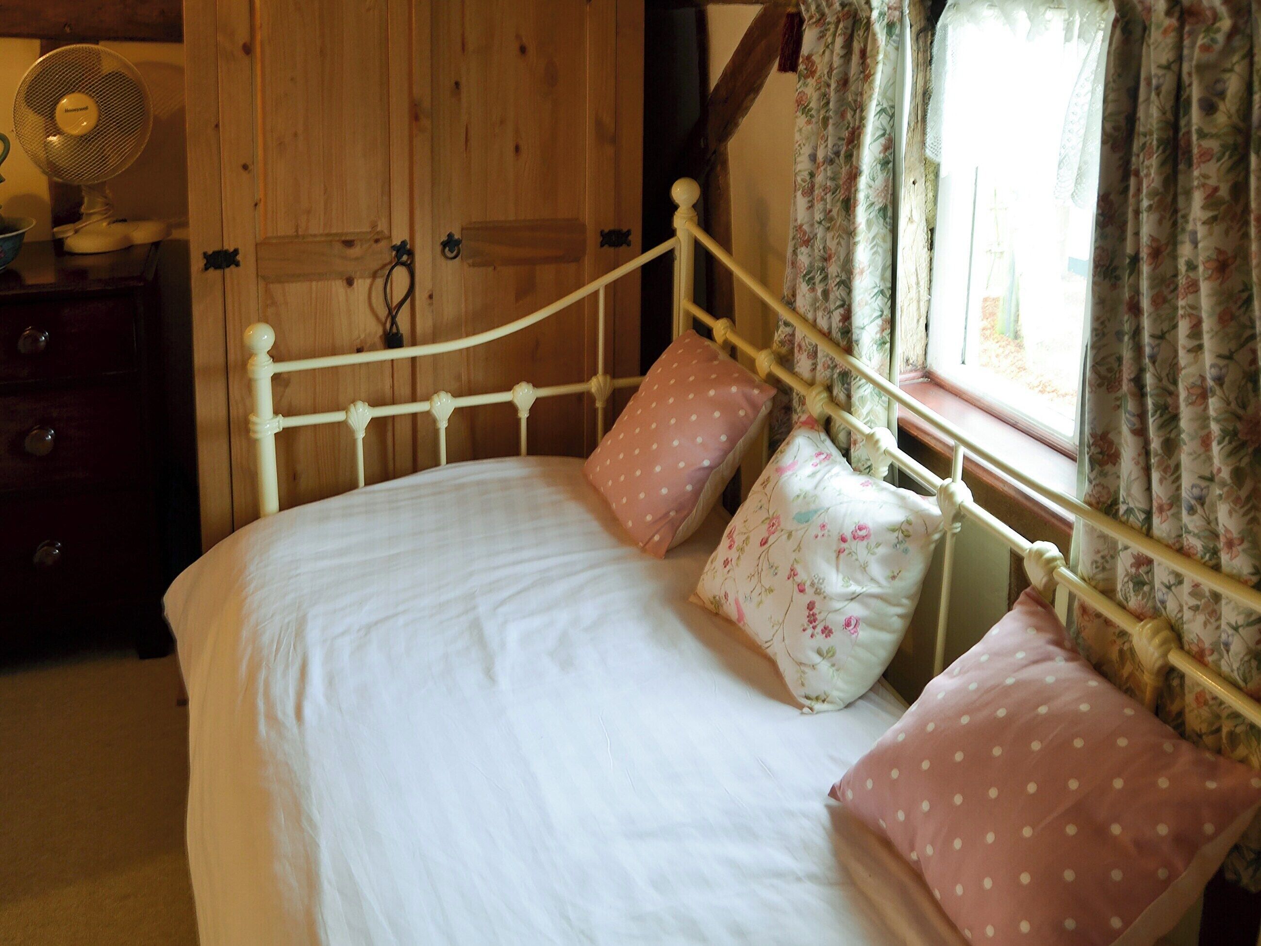 A bedroom with a white bed, floral patterned pillows, a wooden wardrobe, and a window with floral curtains.