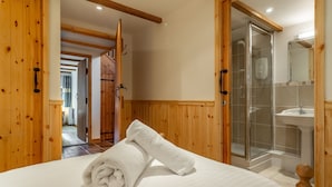 Habitación de hotel con puerta de madera, cama con toalla blanca enrollada, zona de ducha con puerta de cristal y lavamanos.