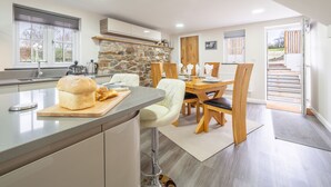 A modern kitchen with a stone accent wall, a dining area with wooden chairs, and a view of the outdoors. - A thoughtfully converted three-bedroom barn conversion, near wonderful beaches. (Chacewater)