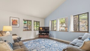 Un salon comprenant un canapé, un téléviseur, un tapis à motifs et de grandes fenêtres donnant sur des arbres et des rochers.