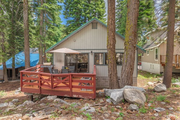 A cabin with a deck, surrounded by trees, and a blue-roofed structure in the background.