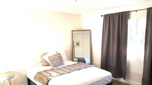 Un dormitorio con cama, mesita de noche, espejo y ventana con cortinas.