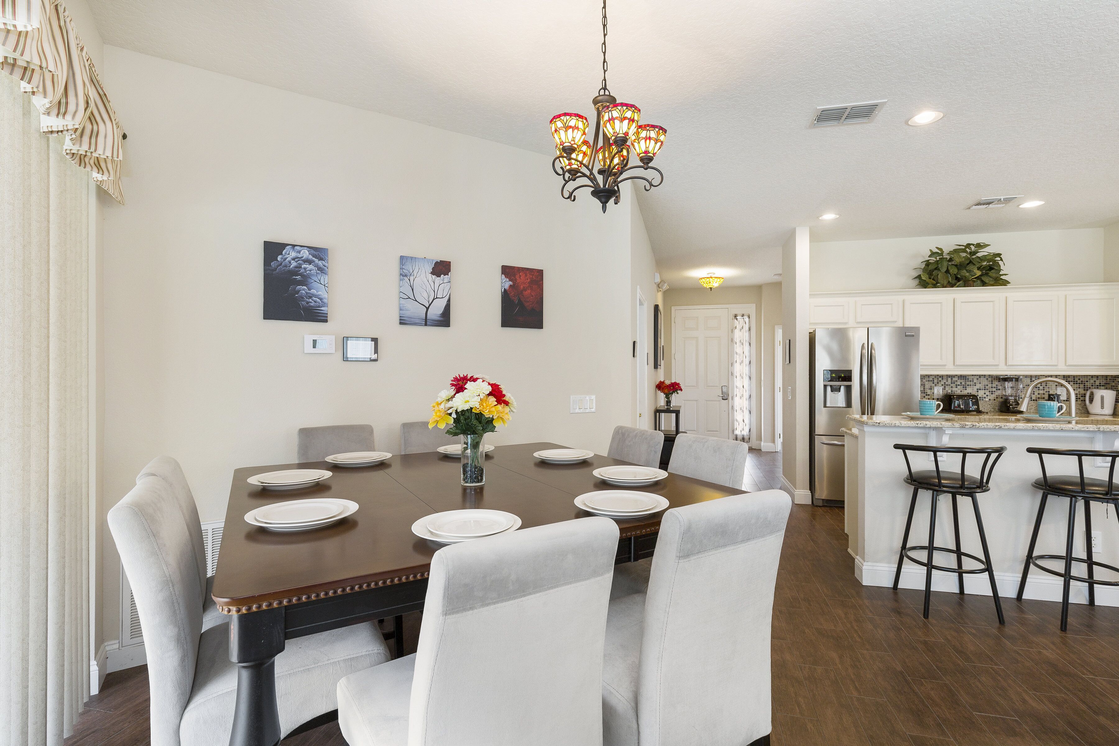 A dining area with a wooden table set for six, surrounded by white chairs. A chandelier hangs from the ceiling, and there are framed pictures on the wall.