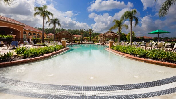 A large pool with a tiled edge, surrounded by lounge chairs and palm trees, with a clear sky above. - Life of Luxury by VillaDirect (Kissimmee)